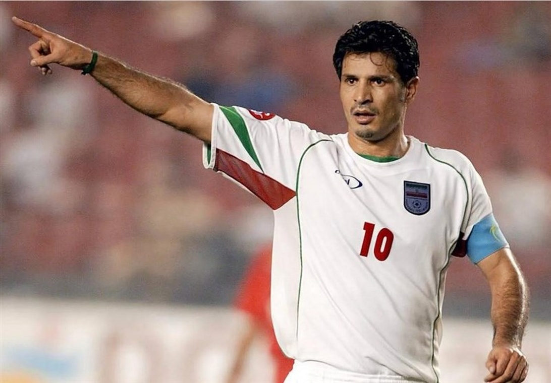 En fotbollsspelare i Irans vita landslagströja med nummer 10 pekar och dirigerar lagkamrater under en match. Han bär kaptensbindeln och hans allvarliga min visar på hans ledarroll.